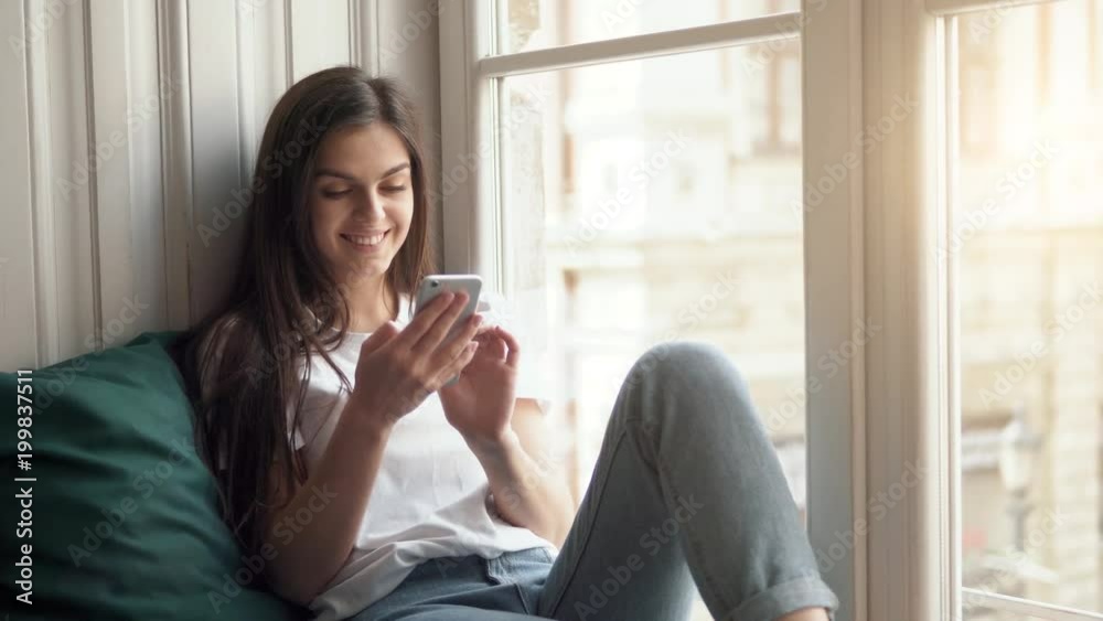 Pretty relaxed girl browsing information on the phone near huge window, using white smartphone happily in modern city flat, wearing white t-shirt and casual blue jeans Pretty relaxed girl browsing information on the phone near huge window, using white smartphone happily in modern city flat, wearing white t-shirt and casual blue jeans