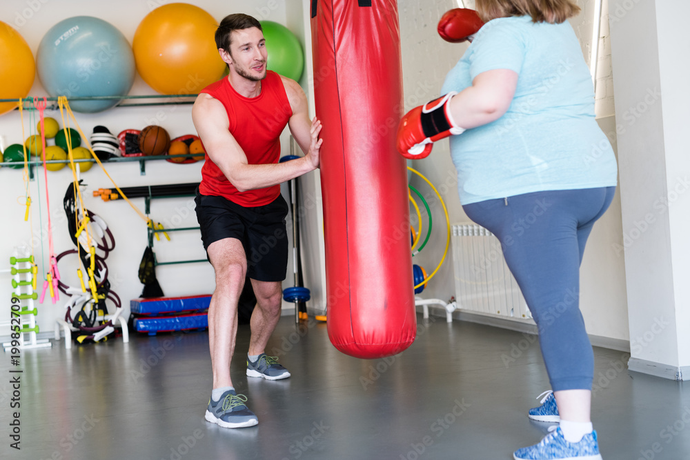 Portrait of handsome fitness instructor holding punching bag while training obese woman to lose weight