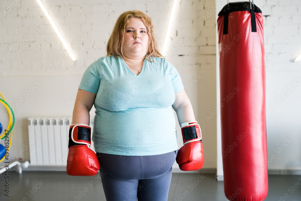 Portrait of obese woman wearing boxing gloves posing next to punching bag and looking at camera, copy space