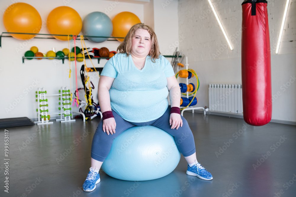 Full length portrait of young obese woman sitting on fitness ball looking at camera, copy space