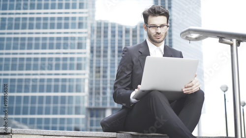 Wallpaper Mural A young prospective business man in a black suit working in the open-air with a laptop. In big city near downtown, sitting on the stairs Torontodigital.ca