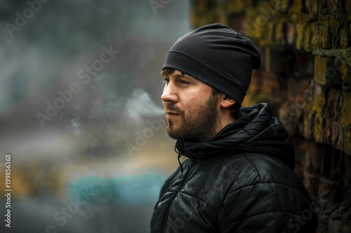 Portrait of a bearded young white guy in the modern hat and black jacket standing near old destroyed red brick wall. Lifestyle. © aleksandr_yu