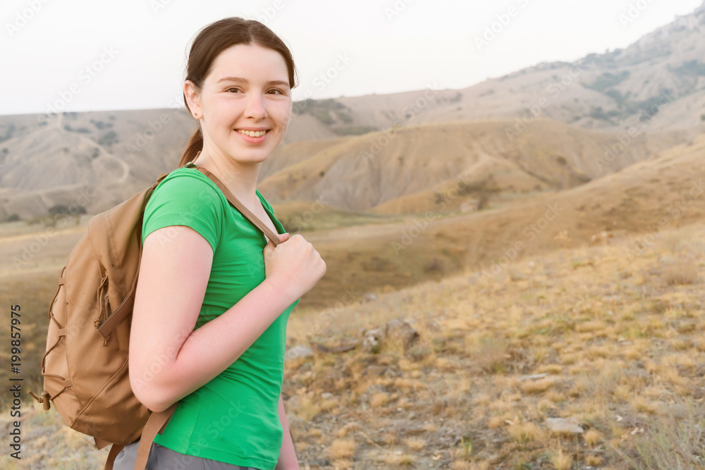 Girl stands sideways to the audience on the belt and holds the strap from the backpack look at the viewer, on the background of rocky steppe