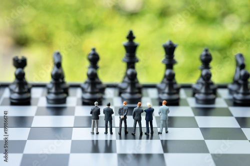 Unity and teamwork in business strategy concept, group of miniature people businessmen collaborate help and work as team, standing on chessboard looking at black chess enemy in far background