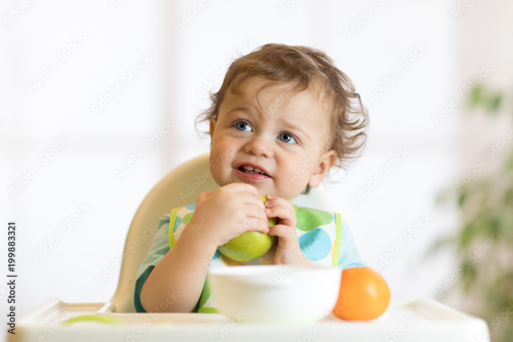 Smiling little kid child baby boy toddler sitting in highchair and eating big green apple fruit portrait indoors Smiling little kid child baby boy toddler sitting in highchair and eating big green apple fruit portrait indoors