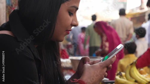 Close up of a pretty woman traveller using her smart phone mobile device during her vacation in Rajasthan Pushkar handheld side profile smile content happy joy touch screen street crowded out of focus