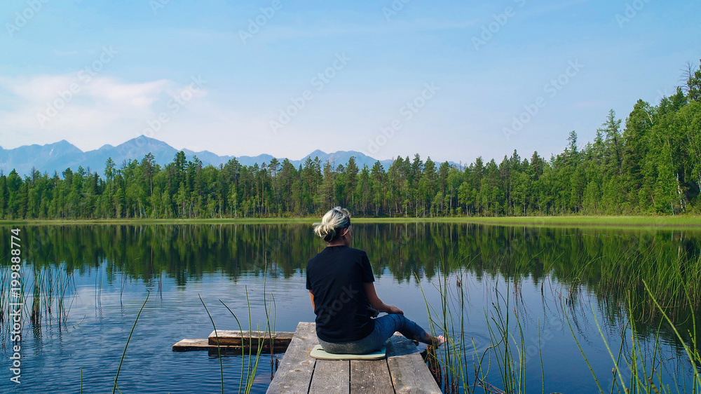 Serenity concept. Pure nature. Lonely Girl sitting on wooden pier and looking at the mountains reflecting in a smooth surface of the lake. Serenity concept. Pure nature. Lonely Girl sitting on wooden pier and looking at the mountains reflecting in a smooth surface of the lake.