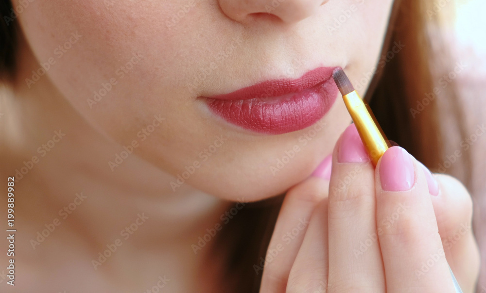 Closeup of a woman's hand with a brush that puts bright lipstick on her lips.