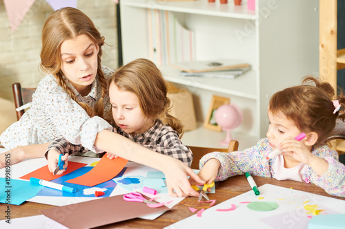 Creative little sisters sitting at wooden table of cozy living room and making handmade present for their mom