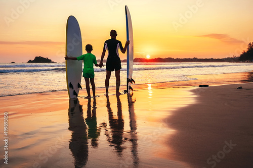 Father with teenager son standing with surfboards on the sandy ocean beach lightened with sunset sun. They smiling and have a conversation. Family active vacation concept
