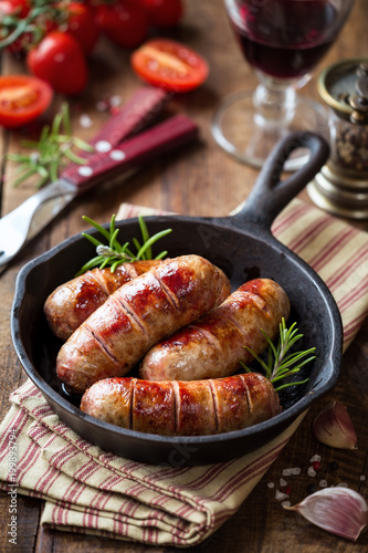 Grilled pork sausages or bangers in cast iron skillet or frying pan with tomatoes, garlic and rosemary