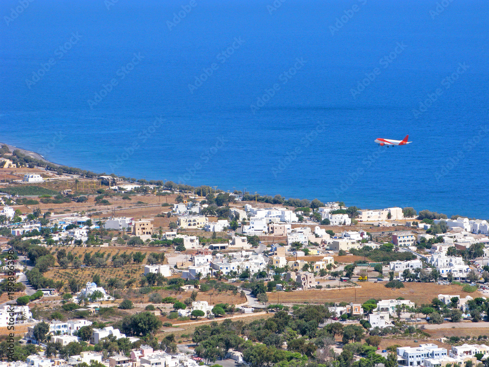 Greece, Cyclades. Summer season. Airplane with vacationers arrives on Santorini island. It flies over the sea. On photo is top view on tourist resort of Kamari and blue sea.