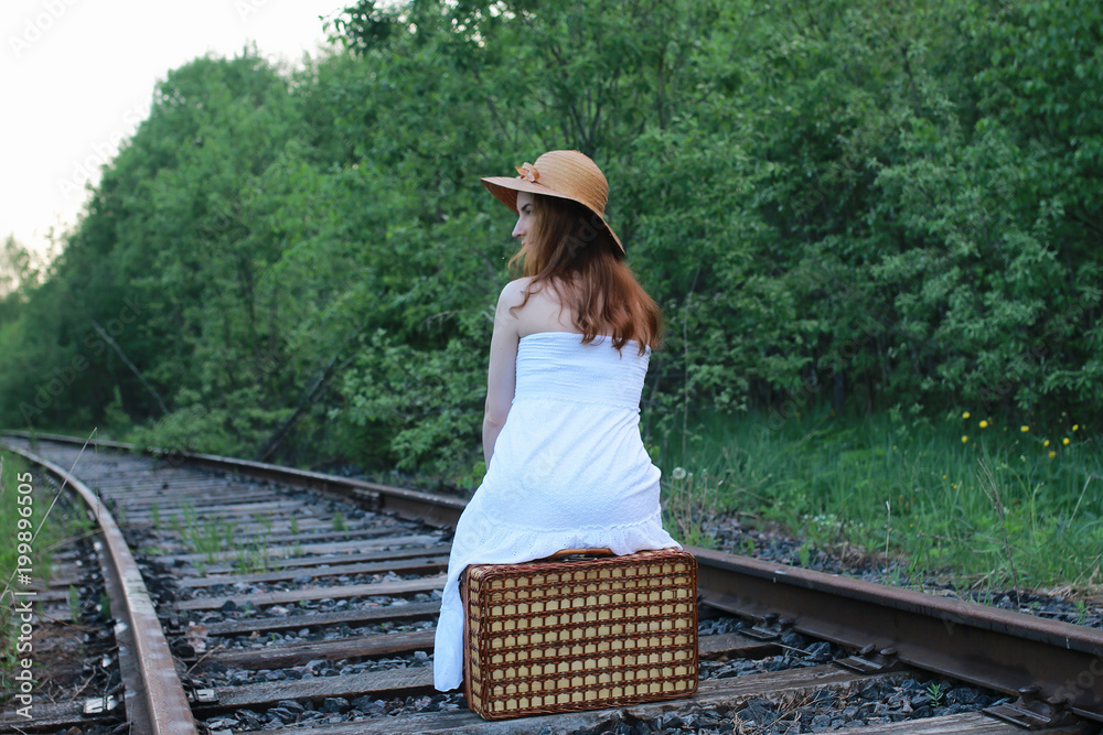 Obraz premium girl in a white sundress and wicker suitcase walking on rails