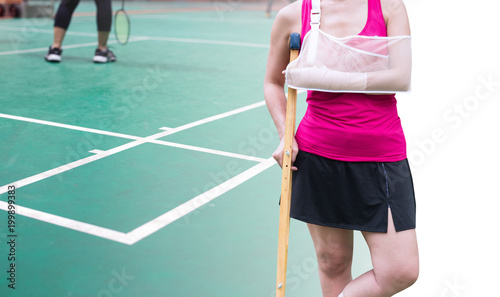 Insurance sport, Injured woman wearing sportswear painful arm with gauze bandage, arm sling and wooden crutches isolated on blurred badminton court with player in game, clipping path included