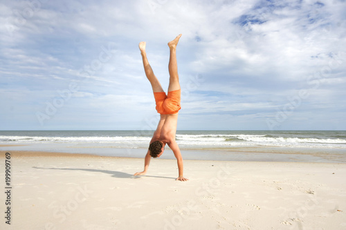 Young man in orange swim suit making hand stand on a sandy beach