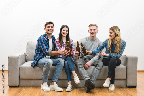 The four happy friends with bottles of beer sit on a white wall background