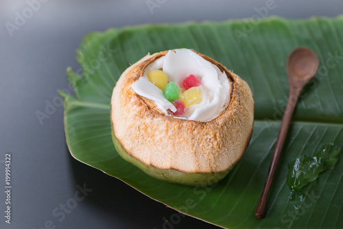 Thai traditional dessert.Tab tim grob on ice cube serve in fresh green coconut shell decorated by coconut meat wooden spoon and banana leaf on black background. photo