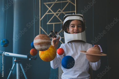 Cute little boy wearing cardboard astronaut helmet flying toy rocket through planets, cardboard spaceship rocket in the background