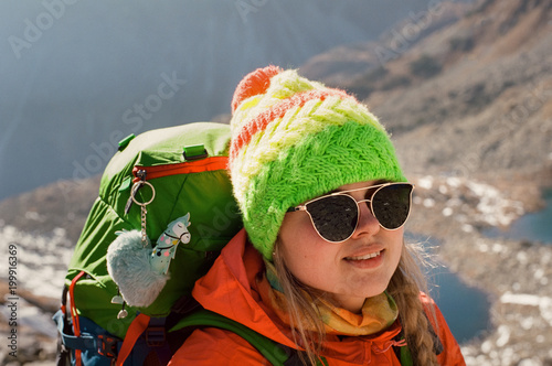 Closeup portrait of happy blonde backpacker in sunglasses and colorful outfit posing on mountain lake background on a sunny day in Slovakia. SShot with film camera and grain photo