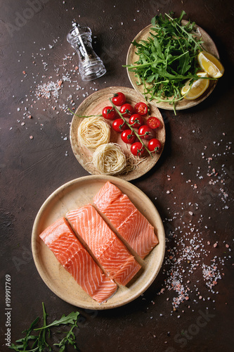 Sliced raw uncooked salmon fillet on ceramic plate with ingredients for dinner, arugula, lemon, salt, pepper, noodles, cherry tomatoes over dark brown texture background. Top view, space.