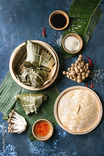 Asian rice piramidal steamed dumplings from rice tapioca flour with meat filling in banana leaves served in bamboo steamer. Ingredients and sauces above over blue texture background. Top view, space.