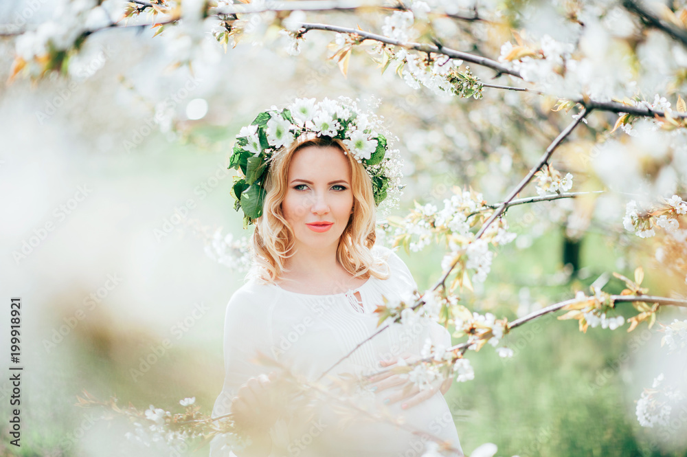 Spring coming! Beautiful young cheerful pregnant woman in wreath flowers on head touching belly while walking in spring tree garden. Beauty People Lifestyle concepts. Happy mom in waiting for baby! Spring coming! Beautiful young cheerful pregnant woman in wreath flowers on head touching belly while walking in spring tree garden. Beauty People Lifestyle concepts. Happy mom in waiting for baby!