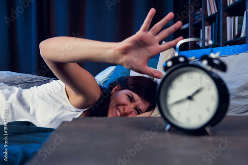 Photo of dissatisfied woman with insomnia stretching arm to alarm clock at night