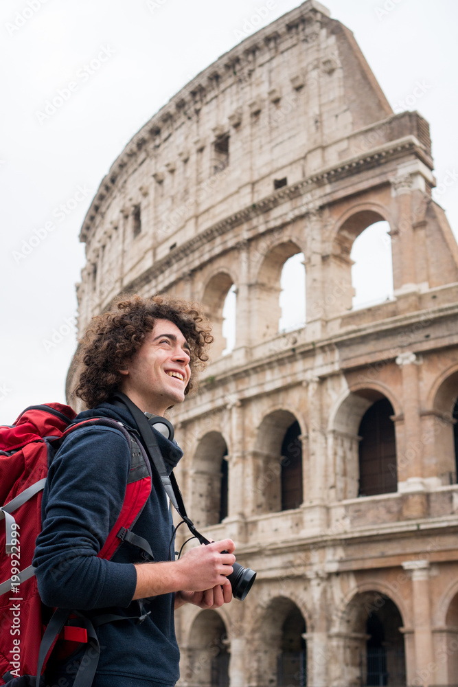Handsome young tourist man with curly hair with a camera and backpack taking pictures of Colosseum in Rome