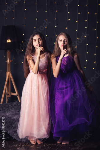 Merry girlfriend. Two girls in pink and purple dresses show shh, in studio on dark background. Copy space.