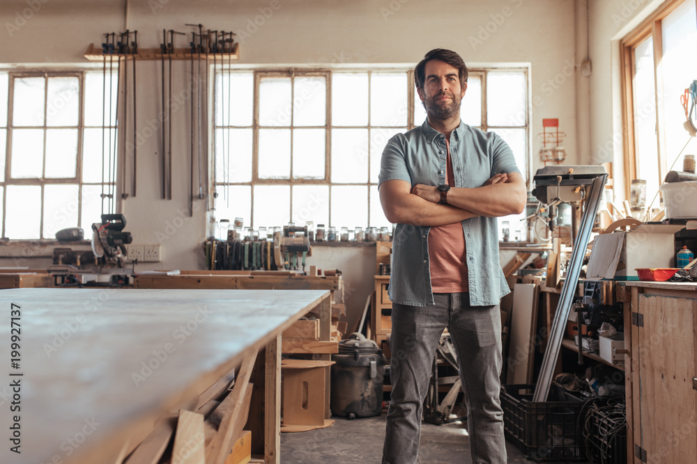 Confident young carpenter standing in his workshop full of tools Confident young carpenter standing in his workshop full of tools
