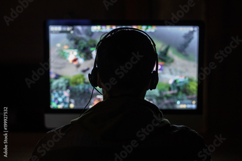 Back view of concentrated young gamer in headphones and glasses using computer for playing game at dark home