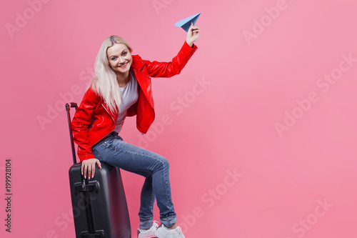 Happy young woman with travel suitcase launches paper airplane. Blonde tourist girl on pink background, concept