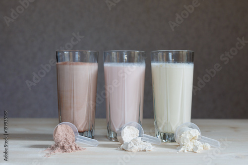 Wallpaper Mural Three milk whey protein shakes in glass cups are arranged in a row with three scoops of protein on a wooden table. Three fresh milk, chocolate, blueberry and banana drinks on table near plastic spoons Torontodigital.ca