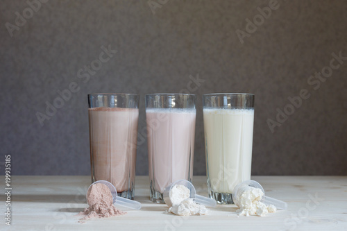 Three milk whey protein shakes in glass cups are arranged in a row with three scoops of protein on a wooden table. Three fresh milk, chocolate, blueberry and banana drinks on table near plastic spoons