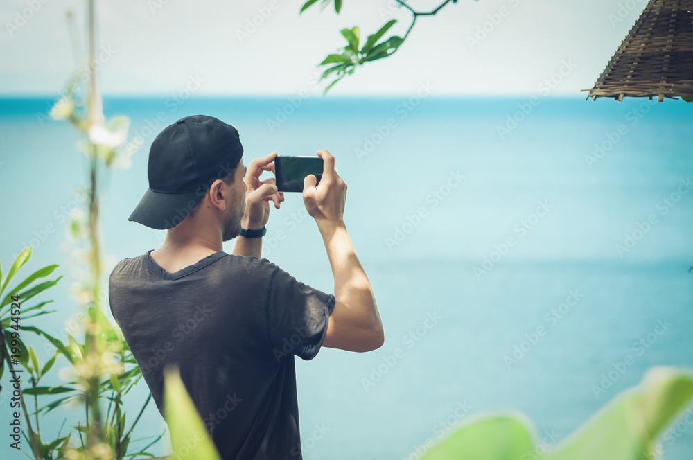 Obraz premium Young man in a cap taking photo of seascape with his cellphone
