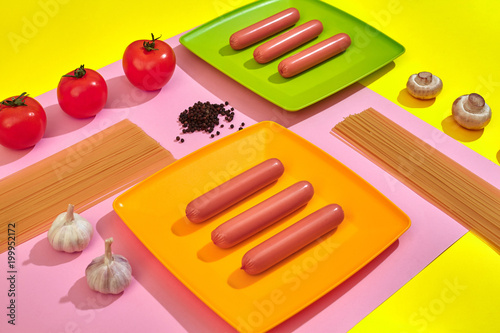 A lot of raw sausages on plate. On pink and yellow background with pasta and vegetables, top view. photo