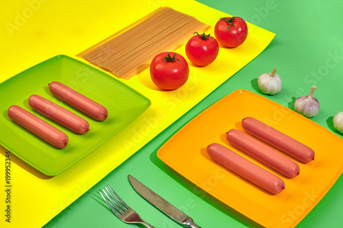 A lot of raw sausages on plate. On green and yellow background with pasta and vegetables, top view. photo