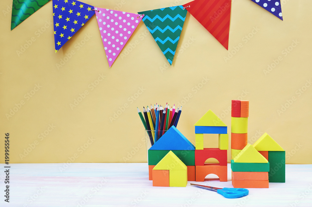 Multicolored festive triangular flags, wooden toy house and glass cup with pencils yellow background