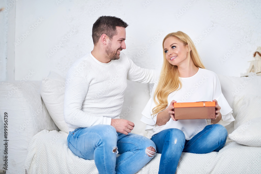 A beautiful happy young couple sitting on the sofa in the living room at christmas and giving presents to each other.