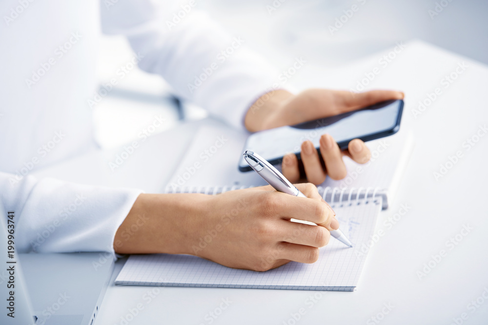 Fototapeta premium Closeup photo of businesswoman's hand holding pen and writing something while using mobile phone