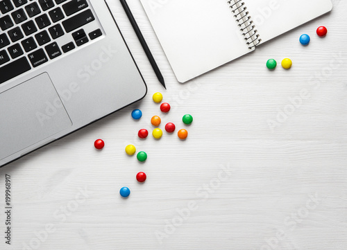 laptop, notebook with a pencil and colored candy on a white wooden background.