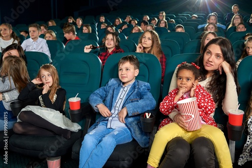 Mother watching intresting movie with children in the cinema hall. Kids are emotional,exited and satisfied. They smiling, laughing, drinking fizzy drinks and eating popcorn.