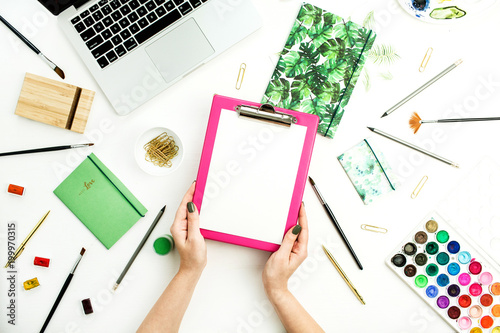 Artistic home office desk workspace. Woman hands holding clipboard. Laptop  notebook  watercolor and stationery. Flat lay  top view blog concept.