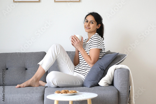 Hübsche dunkelhaarige Frau sitzt auf einer Couch und hält eine Tasse in den Händen lächelt