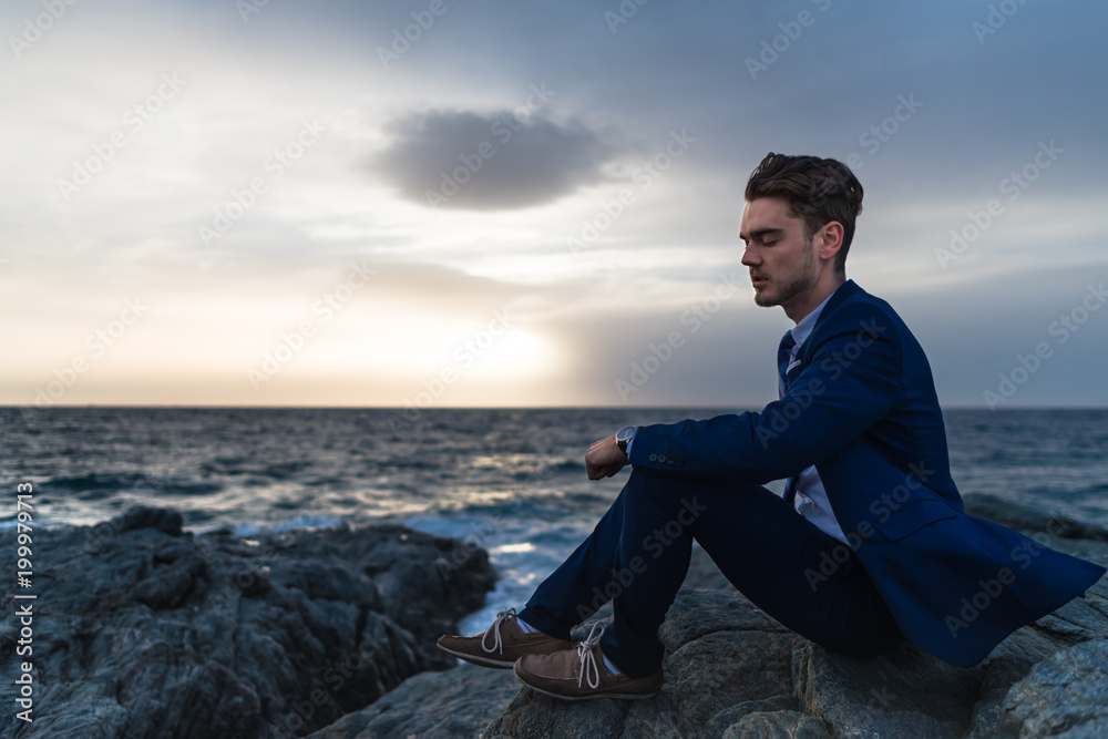 Tired young man in elegant suit sits on the background of the sea and sky. Pensive guy meditating near the ocean in the evening