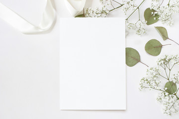 Styled stock photo. Feminine wedding desktop stationery mockup with blank greeting card, baby's breath Gypsophila flowers, dry green eucalyptus leaves, satin ribbon and white background. Empty space