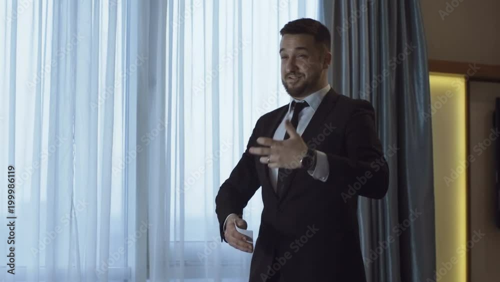 Expressive man in formal suit walking in hotel room with paper notes and training public speech expressively near window. Expressive man in formal suit walking in hotel room with paper notes and training public speech expressively near window.