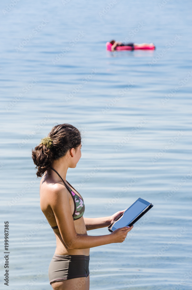 Portrait of young women standing in Lake Constance with digital tablet in her hand and another person on air mattress in the background
