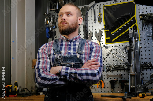 Portrait of a handsome stylish male with beard and haircut wearing a flannel shirt and jeans coverall, standing with crossed arms in a workshop against wall tools.