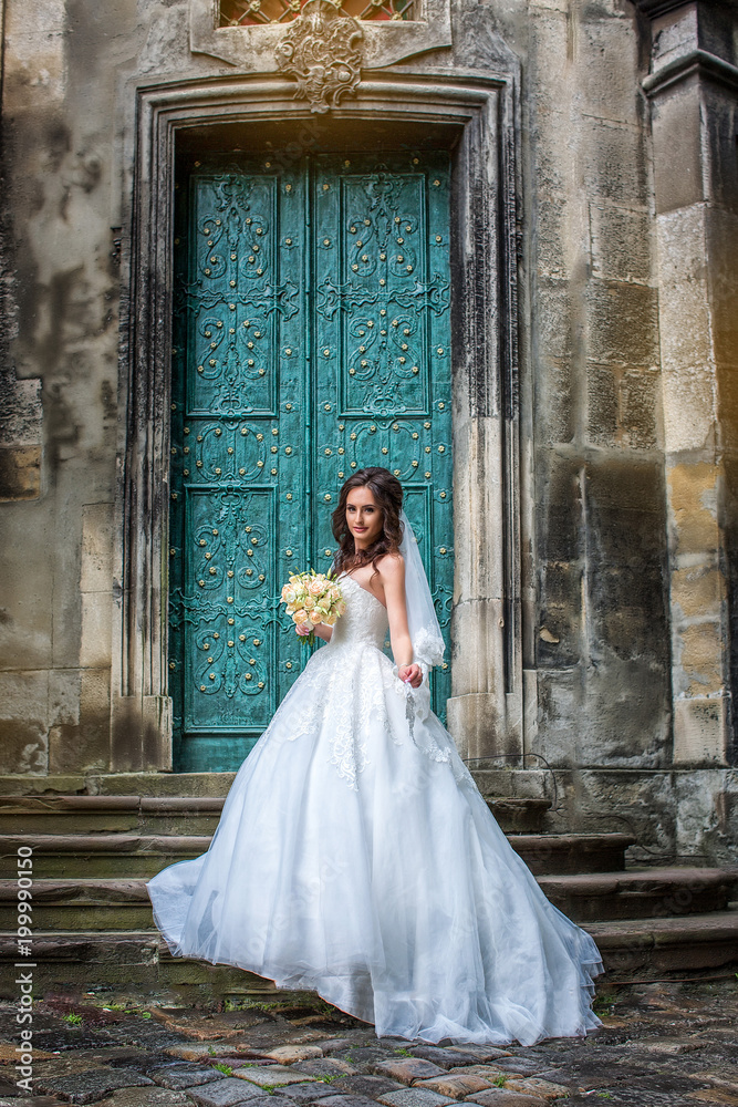 Delightful bride in white dress and veil stands on steps near old green door to cathedral